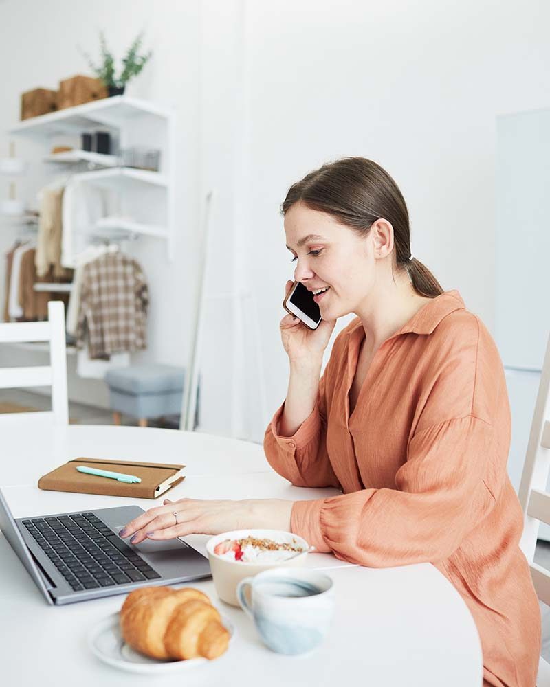 Young businesswoman using laptop during her conversation on mobile phone, while working at the table in the kitchen