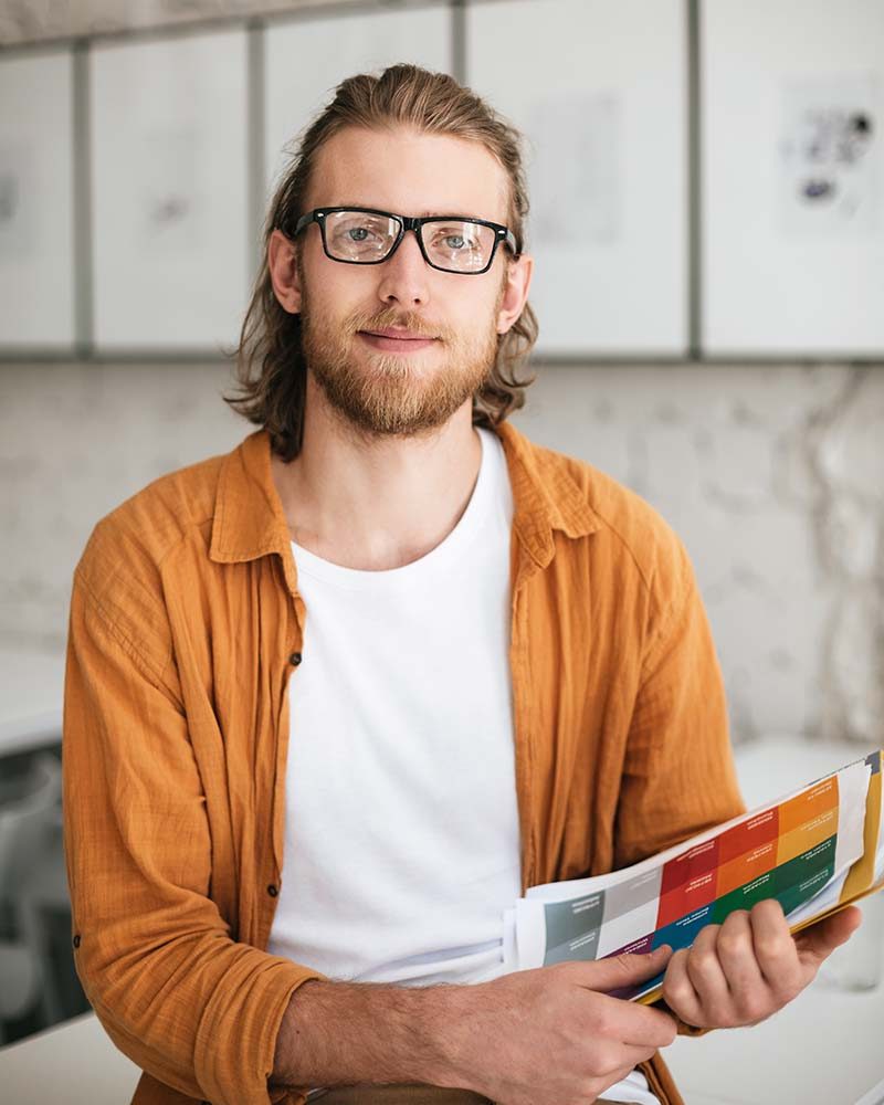 Young man sitting on desk in auditorium with colorful document case in hands. Portrait of smiling boy with blond hair and beard in glasses looking in camera while holding documents in hands
