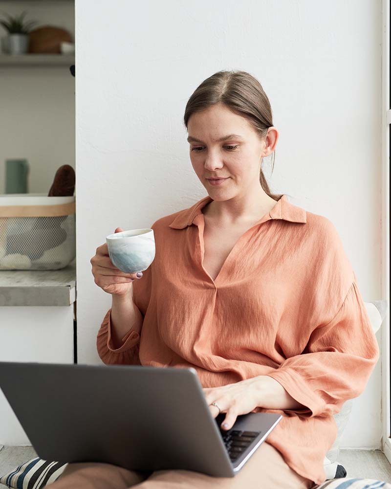 Young woman doing her online work on laptop and drinking coffee while sitting in the room